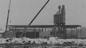 Black and white photo of MCON 1956 factory featuring a large crane in the foreground.